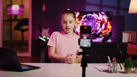 Happy child filming reaction video for gen Z viewers, preparing to look at online videos on laptop, handheld camera shot. Small girl recording reaction to internet clips for social media channelsの写真素材