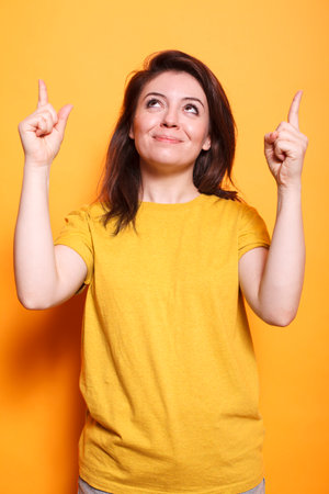 Portrait of joyful brunette lady gesturing with index fingers up and smiling. Smiling caucasian woman using hands to show direction and indicating upwards for photo in studio. Advertisement pose.の写真素材