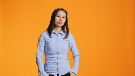 Portrait of carefree asian girl posing with confidence, wearing blue shirt and feeling confident in studio. Pretty filipino person standing over orange background, smiling with elegance.の写真素材