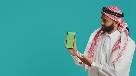 Middle eastern man points at greenscreen display on smartphone, showcasing isolated copyspace template on mobile device screen. Young person in islamic costume shows blank chromakey.の写真素材