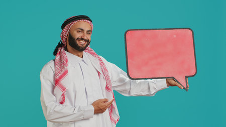 Muslim adult holds speech bubble on camera, creating advertisement for new web commercial. Young person working on marketing industry with empty cardboard icon, isolated billboard sign.の写真素材
