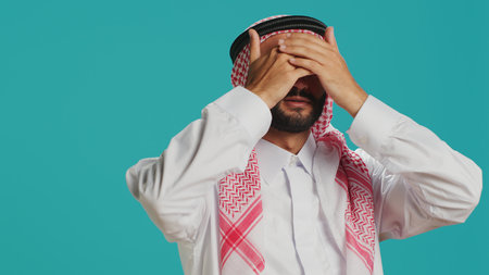 Arab person covers eyes, ears and mouth in studio, presenting three wise monkeys metaphor sign. Middle eastern adult showcasing dont hear, see or speak symbol, muslim costume.の写真素材
