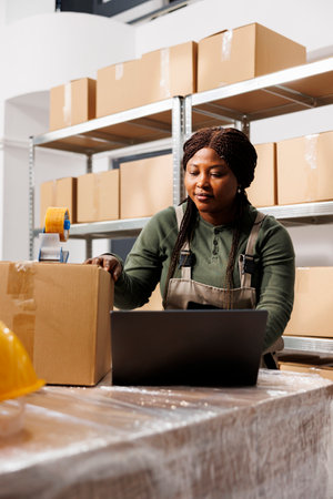 Storehouse worker using carton boxes to pack clients orders, analyzing shipping details on laptop computer in storage room. African american employee wearing industrial overall during inventoryの写真素材