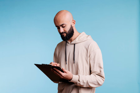 Concentrated arab man planning and writing checklist while holding clipboard. Young focused person wearing casual hoodie checking items in list and taking notes in studioの写真素材