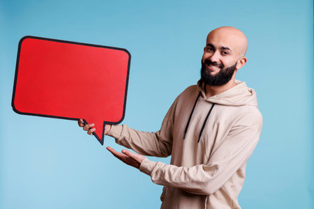 Cheerful arab man promoting new product with comic balloon red banner portrait. Smiling person holding empty dialog bubble and looking at camera with happy facial expressionの写真素材