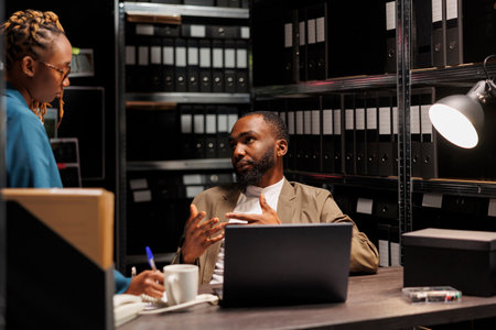 Man and woman investigators brainstorming and discussing crime investigation in dark room full of racks with folders. African american detectives studying reports using archival records and laptopの写真素材
