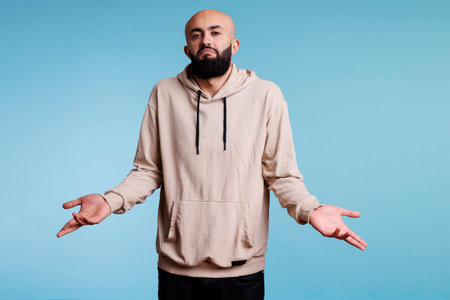 Arab man shrugging shoulders in doubt and confusion while looking a camera. Young person spreading...