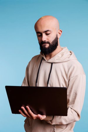 Concentrated arab man reading business report on laptop and typing online message. Focused young person holding portable computer, using software apps and browsing internetの写真素材