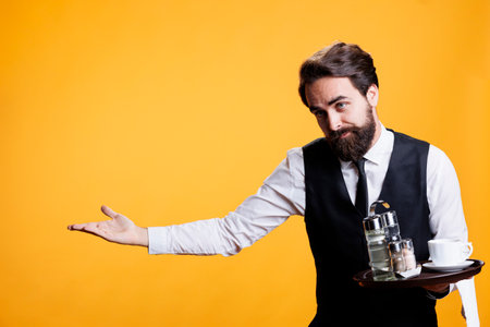 Friendly gracious waiter makes way for people to enter restaurant, providing great service and amazing food at five star place. Young adult with suit and tie posing against yellow background.の写真素材