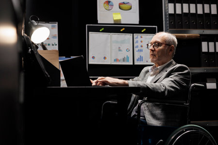 Experienced business manager executive in wheelchair at office desk inspecting quarterly company results analysis data on laptop. Senior with disability on file cabinet office filled with flowchartsの写真素材