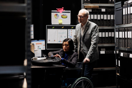 Bookkeeping businesswoman with disability in wheelchair and coworker working in administrative office surrounded by flowcharts. Consultancy bureaucratic workplace filled with business foldersの写真素材