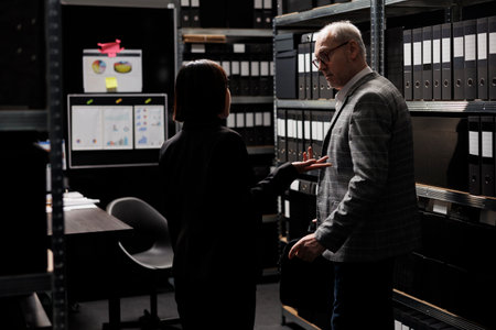 Accountancy executives discussing quarterly profit revenue, analysing performance data. Asian woman and senior businessman in bureaucratic workplace surrounded by documentation folders in cabinetsの写真素材