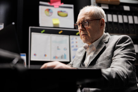 Old business manager executive sitting at office desk before consulting meeting, inspecting quarterly company results analysis data paperwork on laptop. Bureaucratic workplace filled with flowchartsの写真素材