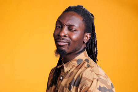 Smiling playful african american man winking at camera, fooling around over yellow background. Portrait of positive young adult having fun doing eye wink in studio, enjoying free time.の写真素材