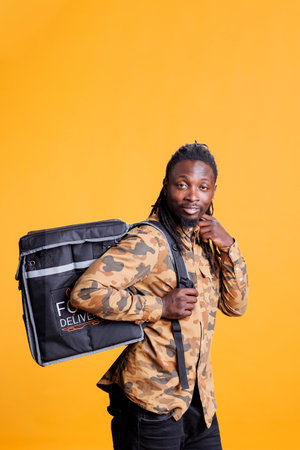 Cheerful courier holding food delivery thermal backpack, standing in studio over yellow background. African american restaurant worker deliverying fast food order to customers during lunch timeの写真素材