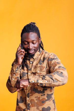 African american man checking watchwrist, talking with remote friend using mobile phone in studio. Excited successful young adult discussing about lifestyle, standing over yellow backgroundの写真素材