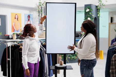 Woman showcasing latest shoe collection to customer on smart display in clothing store. Shopping mall employee using blank whiteboard to assist shopper with selecting trendy stilettoの写真素材