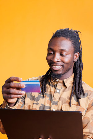 African american man using credit card to shop online using smartphone fashion clothing app, ordering stylish clothes. Young adult paying products on digital store in studio over yellow backgroundの写真素材
