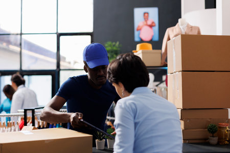 African american courier showing logistics report to worker, discussing customer shipping details in boutique. Employee preparing packages for delivery, working with online orders in shopping centreの写真素材