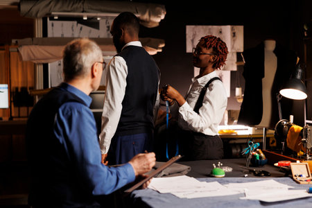 African american gentleman getting measurements taken by professional suitmakers in dark studio workspace. Masterful tailors looking for ideal client proportions for upcoming custom made suitの写真素材