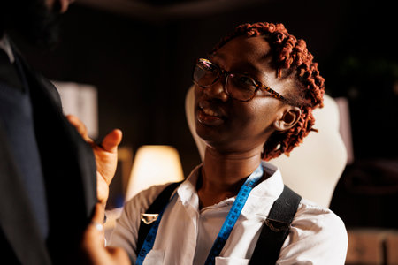 Close up of african american tailor helping customer try on bespoken sartorial piece after finishing needlework comission. Seamstress manufacturing elegant business attire in black owned atelier shopの写真素材