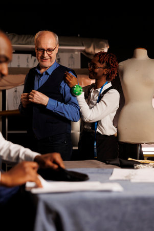 Cheerful meticulous seamstress doing precise additional needlework needed on sartorial piece worn by elderly client. Skilled african american tailor helping customer with bespoke suit comissionの写真素材