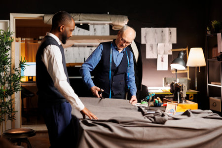 Meticulous elderly master tailor cutting fabric sheet for upcoming bespoke luxury fashion collection. Senior suitmaker showing african american apprentice expert craftmanshipの写真素材