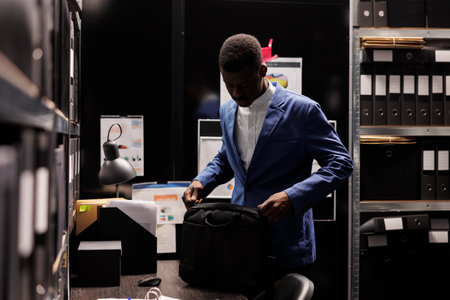 African american detective preparing his backpack, leaving job late at night in arhive room. Police officer working overtime at criminal case, analyzing crime scene evidence. Criminology conceptの写真素材
