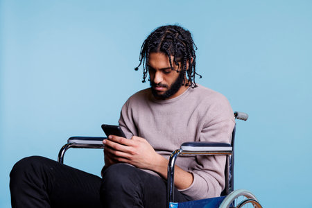 Arab man sitting in wheelchair while scrolling social media on smartphone. Young person with physical impairment sending online message and browsing internet on mobile phoneの写真素材