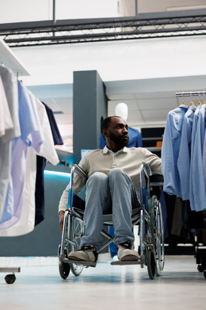 Clothing store customer with physical disability choosing formal shirt and exploring apparel rack. African american man in wheelchair shopping for outfit in menswear boutiqueの写真素材