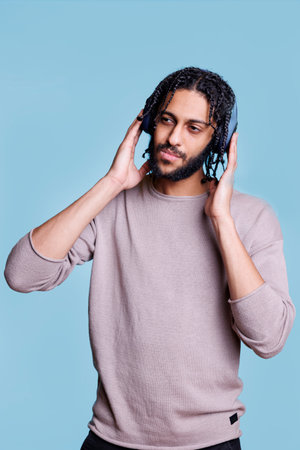 Arab man listening to song, touching wireless headphones on head and looking away with pensive...