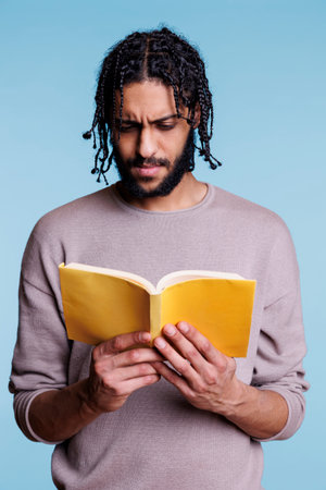 Thoughtful focused arab student reading novel book with yellow cover. Concentrated pensive young adult man studying literature while holding softcover paperback open textbookの写真素材