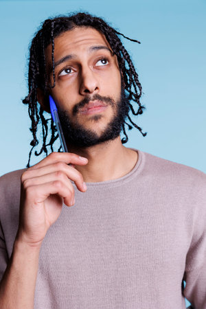 Pensive arab man thinking while looking away with puzzled facial expression and touching cheek with pen. Thoughtful person making decision and planning with uncertain emotionsの写真素材