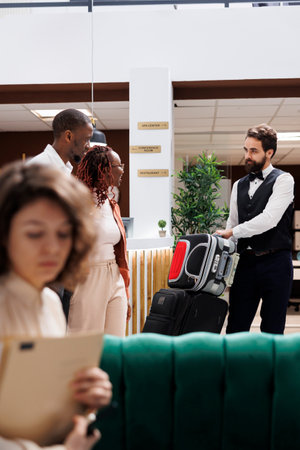 Male employee helping clients with bags, carrying luggage for people after doing check in procedure. Young couple receiving assistance from luxury resort bellboy at reception desk.の写真素材
