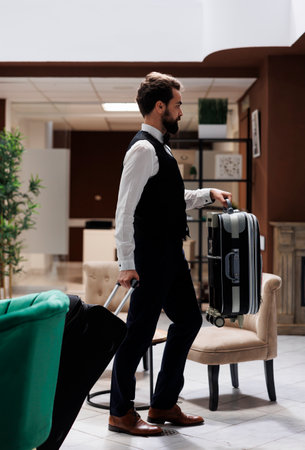Male hotel concierge carrying bags to help guests in reception area, moving suitcases and tolleys for tourists checking in at luxury resort. Young elegant bellboy giving assistance for people.の写真素材