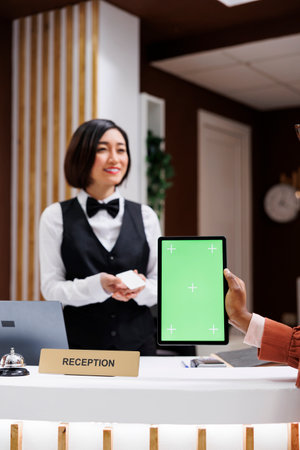 Woman holding tablet with greenscreen at front desk, receiving room access card from asian receptionist. Young person using isolated mockup display with chromakey at hotel reception.の写真素材