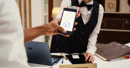 Close up of african american hotel visitor using smartphone banking app to buy holiday package deal upgrade. Receptionist using POS payment terminal to validate credit card purchase during check inの写真素材