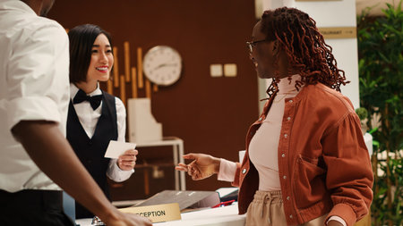 Happy receptionist handing relaxed smiling newlywed couple room access key card during check in process. Cheerful tourists and professional hotel staff chatting in resort lobbyの写真素材