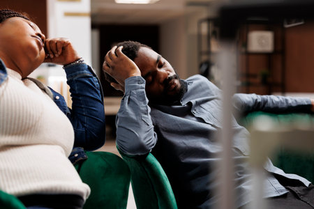 Tired unhappy African American family couple sleep in hotel lobby having jet lag symptoms, tourists feeling sleepy and exhausted after long flight. Air travel and sleep deprivationの写真素材