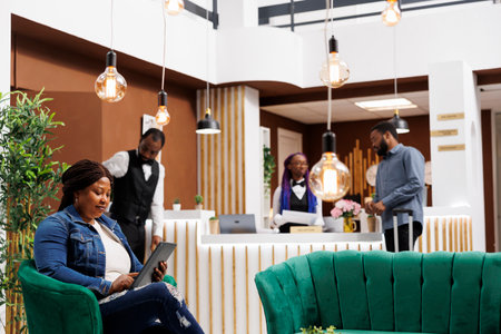 African American woman sitting in lobby with tablet using digital technology to check-in into hotel, tourist holding electronic device browsing internet while resting at resort foyerの写真素材