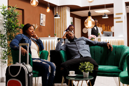 Exhausted tourists resting in hotel lobby, waiting for check-in, sleeping after long day of traveling. Tired African American couple of travelers fall asleep, taking nap, adapting to new time zoneの写真素材