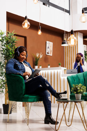 Relaxed African American woman tourist wearing wireless headphones using digital tablet while relaxing in cozy hotel rest zone, watching movie online and drinking coffee while waiting for check-inの写真素材