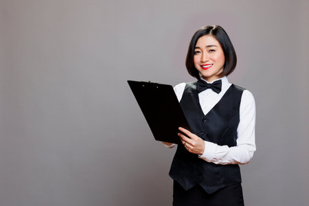 Attractive smiling asian waitress writing on a clipboard and looking at camera with positive facial expression. Cheerful young receptionist in uniform taking notes while posing in studio portraitの写真素材