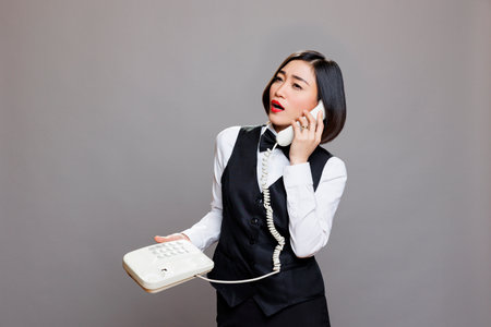 Asian woman receptionist in uniform talking on landline phone, giving instruction to restaurant staff. Confident restaurant worker answering customer call using retro telephone in studioの写真素材