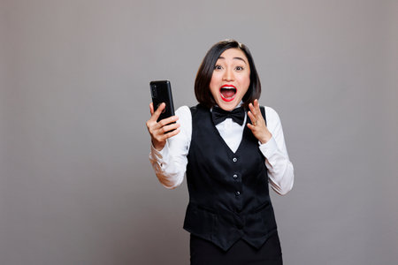 Excited asian waitress holding smartphone and receiving good news while looking at camera with...