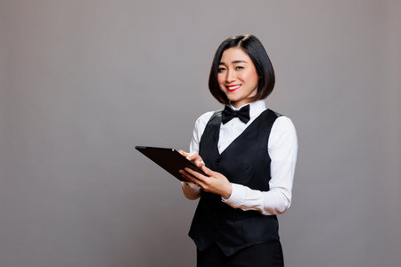 Smiling asian woman receptionist using digital tablet and looking at camera with positive facial expression. Catering service waitress in uniform standing with portable device portraitの写真素材