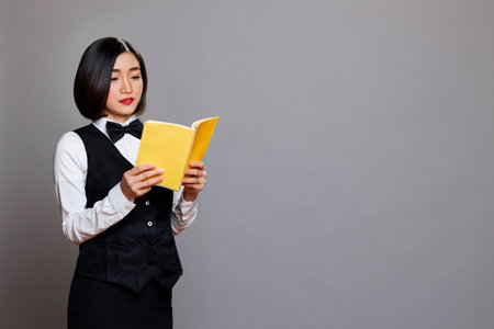 Concentrated asian woman receptionist reading book while taking break. Catering service young waitress wearing professional uniform learning cafe menu with yellow blank coverの写真素材