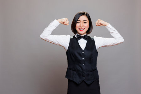 Smiling asian waitress in uniform showing arms muscles and looking at camera with joyful facial expression. Cheerful receptionist displaying biceps, showcasing strength and power portraitの写真素材