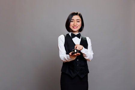 Smiling asian restaurant receptionist in uniform ringing dining bell while posing and looking at...