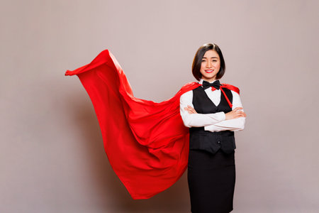 Confident smiling waitress wearing superhero red cape, showing strength and power. Catering service cheerful woman worker in uniform and superwoman cloak standing with folded hands portraitの写真素材